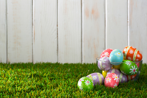 This is a full frame photo of a lot of real bright colorful speckled decorated Easter eggs on a bed of green grass with a white fence in the background.. This is a great image for an esater egg hunt with no people in it.